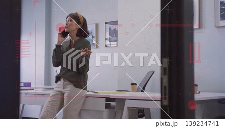 Leaning Asian woman holding smartphone and gesturing at office desk with laptop, papers and coffee Leaning Asian woman holding smartphone and gesturing at office desk with laptop, papers and coffee 139234741