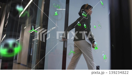 Walking woman in sweater carrying silver laptop through glass door office corridor, green AR icons Walking woman in sweater carrying silver laptop through glass door office corridor, green AR icons 139234822