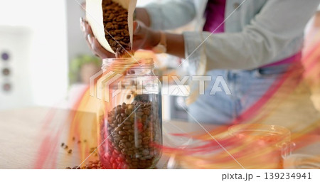 Woman in denim jacket pouring coffee beans from paper bag into glass jar on kitchen countertop 139234941