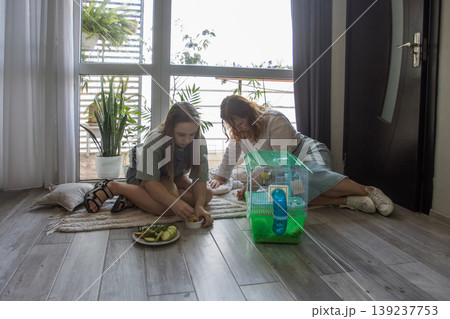 Two girls share snacks and play with a pet on a rug inside a bright room with large windows 139237753