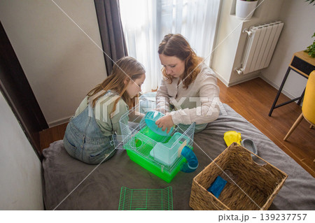 A girl and a woman work together to arrange a green hamster cage on a bed. 139237757