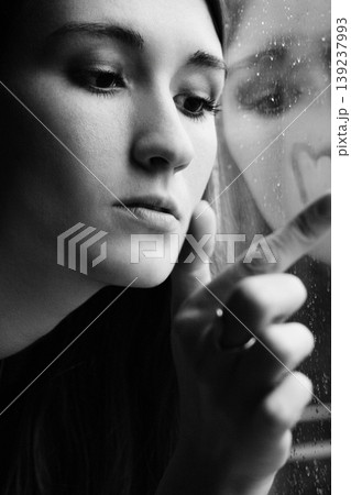 Close-up of a pensive woman drawing a heart on a foggy window Close-up of a pensive woman drawing a heart on a foggy window 139237993
