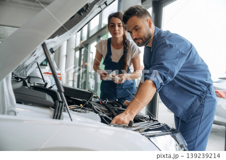 View from the side, hood is opened. Auto repair shop employees are working together. Man and woman View from the side, hood is opened. Auto repair shop employees are working together. Man and woman 139239214