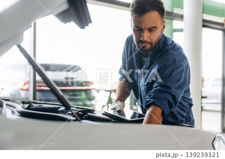 Black digital tablet, hood is opened, fixing the automobile. Male mechanic is working in the modern car workshop 139239321