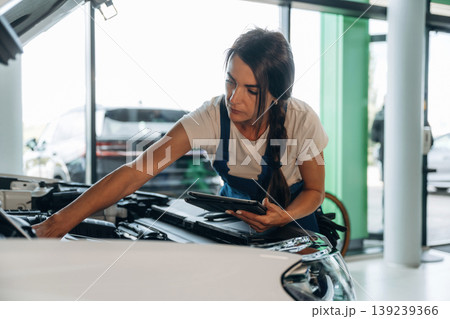 Against window, hood is opened, with digital tablet. Female mechanic is working in the modern car workshop Against window, hood is opened, with digital tablet. Female mechanic is working in the modern car workshop 139239366
