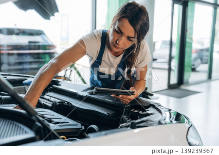 Against window, hood is opened, with digital tablet. Female mechanic is working in the modern car workshop 139239367