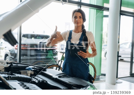 Against window, hood is opened, with digital tablet. Female mechanic is working in the modern car workshop Against window, hood is opened, with digital tablet. Female mechanic is working in the modern car workshop 139239369