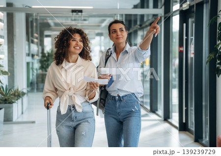 Tourists with suitcase. Two women are indoors in the modern hall of building at daytime Tourists with suitcase. Two women are indoors in the modern hall of building at daytime 139239729