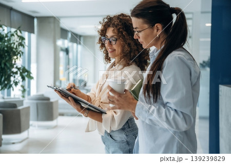 Business partners are together. Two women are indoors in the modern hall of building at daytime Business partners are together. Two women are indoors in the modern hall of building at daytime 139239829