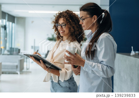 Writing the information into notepad. Two women are indoors in the modern hall of building at daytime Writing the information into notepad. Two women are indoors in the modern hall of building at daytime 139239833