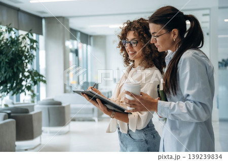 Writing the information into notepad. Two women are indoors in the modern hall of building at daytime 139239834