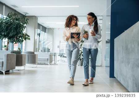 Writing the information into notepad. Two women are indoors in the modern hall of building at daytime Writing the information into notepad. Two women are indoors in the modern hall of building at daytime 139239835