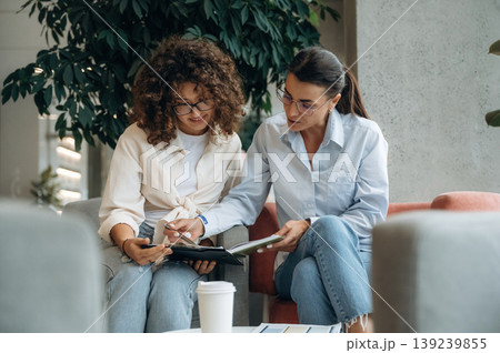 Sitting on the sofa with notepad. Two women are indoors in the modern hall of building at daytime Sitting on the sofa with notepad. Two women are indoors in the modern hall of building at daytime 139239855