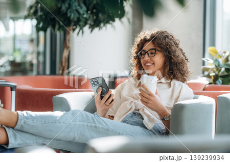 On the sofa, relaxing, using smartphone. Beautiful woman is in airport hall with suitcase 139239934