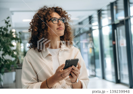 Smartphone is in hands. Pretty woman in casual clothes is in the modern building indoors 139239948
