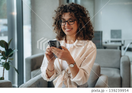 Smartphone is in hands. Pretty woman in casual clothes is in the modern building indoors Smartphone is in hands. Pretty woman in casual clothes is in the modern building indoors 139239954