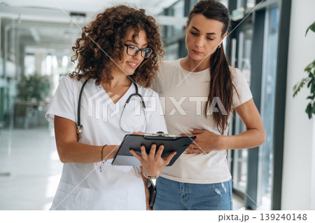 Consultation with doctor. Two women are indoors in the modern hall of building at daytime Consultation with doctor. Two women are indoors in the modern hall of building at daytime 139240148