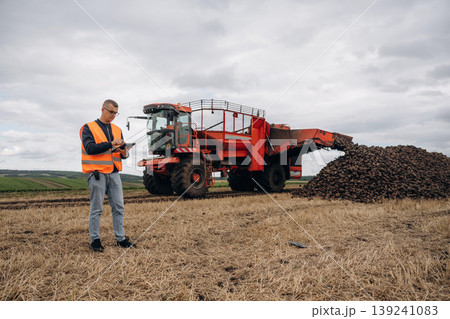 Digital tablet is in hands. Farmer is working in the agricultural field. Against beet harvester Digital tablet is in hands. Farmer is working in the agricultural field. Against beet harvester 139241083