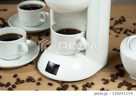 White drip coffee maker with a full cup of fresh coffee and scattered beans. White drip coffee maker with a full cup of fresh coffee and scattered beans. 139241154