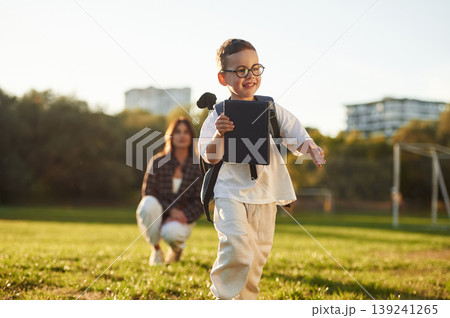 Walking forward, pupil is with book and backpack. Mother and son together in an open field during daytime 139241265