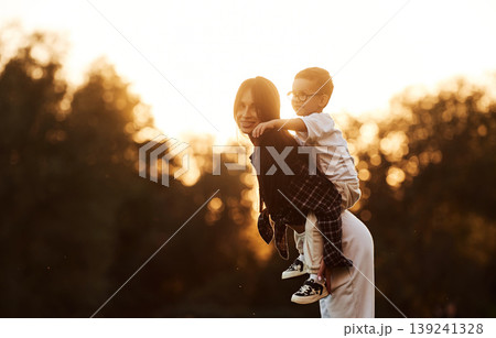 Trees illuminated by evening sunlight, background. Mother and son together in an open field during daytime 139241328