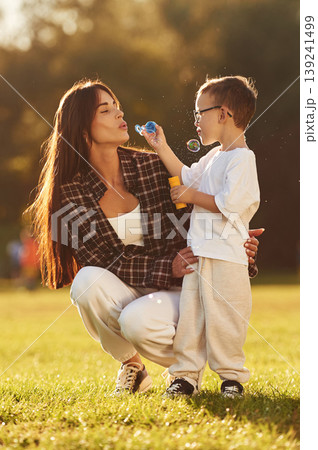 Front view, blowing bubbles. Mother and son together in an open field during daytime Front view, blowing bubbles. Mother and son together in an open field during daytime 139241499