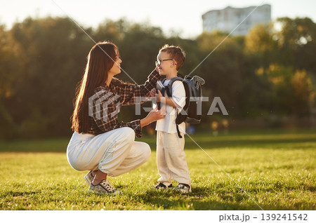 Getting ready for school. With book and backpack. Mother and son together in an open field during daytime Getting ready for school. With book and backpack. Mother and son together in an open field during daytime 139241542