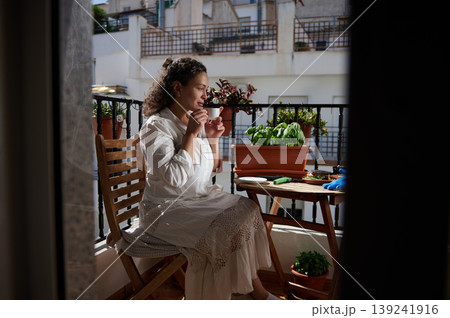 Woman Drinking Coffee on Sunny Balcony Surrounded by Potted Herbs and Plants Woman Drinking Coffee on Sunny Balcony Surrounded by Potted Herbs and Plants 139241916