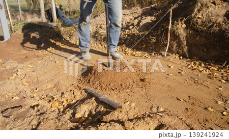 worker leveling soil with tools outdoors, person spreading loose soil on sunny site using garden tools, landscaper using shovel and rake to prepare ground for planting or construction 139241924
