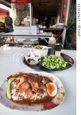 A Thai street food staple: Khao Moo Daeng. Slices of roasted red pork and crispy belly rest on jasmine rice, drenched in a rich, five-spice gravy. Accompanied by a soft-boiled egg, fresh cucumbers, A Thai street food staple: Khao Moo Daeng. Slices of roasted red pork and crispy belly rest on jasmine rice, drenched in a rich, five-spice gravy. Accompanied by a soft-boiled egg, fresh cucumbers, 139243019