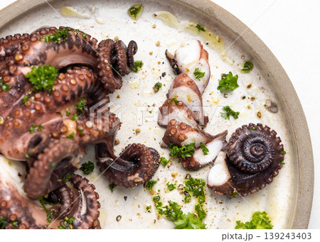 Boiled octopus tentacles and sliced pieces on ceramic plate top view. Olive oil and parsley dressing 139243403