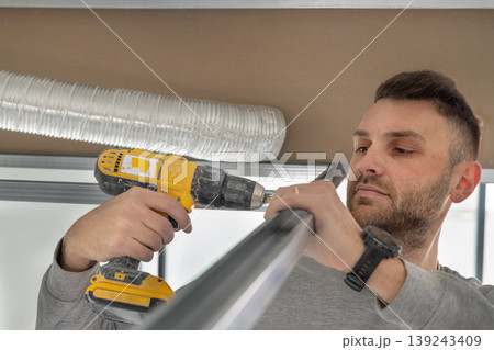 Construction worker using drill on metal frame for drywall suspended ceiling in apartment renovation Construction worker using drill on metal frame for drywall suspended ceiling in apartment renovation 139243409