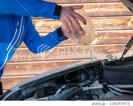Man pouring water from plastic bottle into car radiator. Driver performing routine vehicle maintenance under hood. Engine fluid refill process for automotive repair and transport care. Man pouring water from plastic bottle into car radiator. Driver performing routine vehicle maintenance under hood. Engine fluid refill process for automotive repair and transport care. 139243473