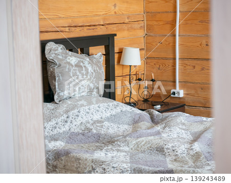 Cozy bedroom corner with wooden wall and blanket in rustic house. Comfortable bed with pillow and bedside lamp in cottage interior. Design of traditional wooden home sleeping area. Cozy bedroom corner with wooden wall and blanket in rustic house. Comfortable bed with pillow and bedside lamp in cottage interior. Design of traditional wooden home sleeping area. 139243489