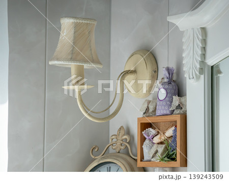 Vintage lamp mounted on wall with decorative shelf containing lavender sachets and flowers. Interior design detail with antique style elements for cozy home living and cottage decor. Vintage lamp mounted on wall with decorative shelf containing lavender sachets and flowers. Interior design detail with antique style elements for cozy home living and cottage decor. 139243509