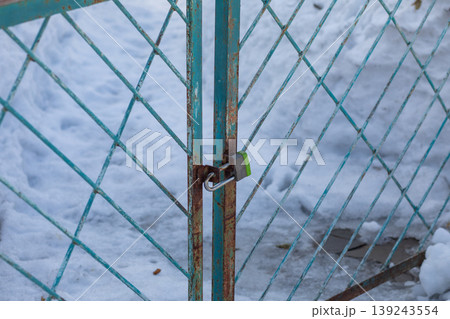Metal gate with rusty chain link mesh locked by padlock. Secured entrance door in winter season with snow background. Concept of safety, protection, restriction, private area closure. Metal gate with rusty chain link mesh locked by padlock. Secured entrance door in winter season with snow background. Concept of safety, protection, restriction, private area closure. 139243554