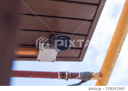 Security camera mounted on brown ceiling under roof. Surveillance system for property protection and safety monitoring. Digital video equipment for prevention of burglary and crime control. Security camera mounted on brown ceiling under roof. Surveillance system for property protection and safety monitoring. Digital video equipment for prevention of burglary and crime control. 139243565