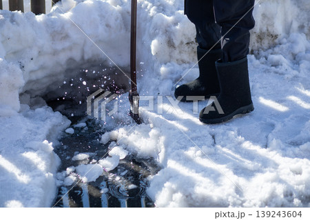 Man breaking ice with ice pick on sidewalk. Winter street snow removal and maintenance. Worker clearing frozen path surface. Safe movement during cold season and icy road conditions. 139243604