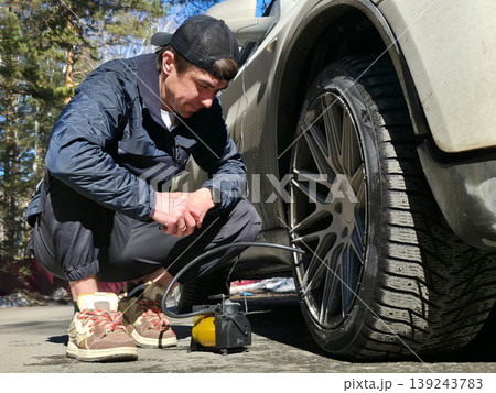 Man inflating car tire with portable electric pump. Driver checking air pressure in wheel on road. Automotive maintenance and vehicle emergency repair procedure. Diagnostic roadside help. Man inflating car tire with portable electric pump. Driver checking air pressure in wheel on road. Automotive maintenance and vehicle emergency repair procedure. Diagnostic roadside help. 139243783