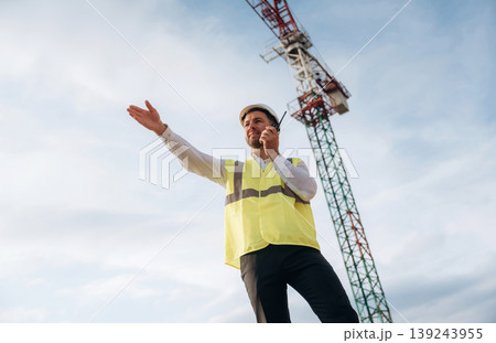 Controlling the process, with walkie talkie. Construction site worker is against building crane and sky background Controlling the process, with walkie talkie. Construction site worker is against building crane and sky background 139243955
