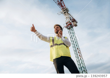 Controlling the process, with walkie talkie. Construction site worker is against building crane and sky background Controlling the process, with walkie talkie. Construction site worker is against building crane and sky background 139243957