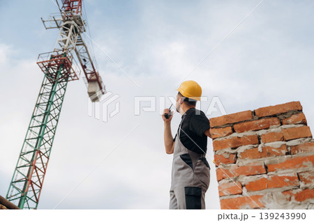 Talking by walkie talkie. A builder is working at the construction site at daytime Talking by walkie talkie. A builder is working at the construction site at daytime 139243990