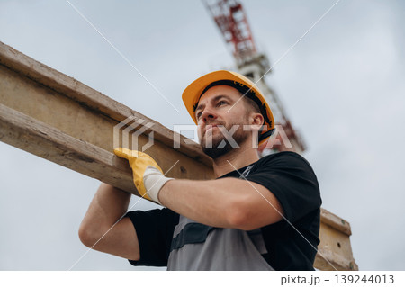 A construction worker is moving the wooden plank. In the yellow hard hat and in gloves A construction worker is moving the wooden plank. In the yellow hard hat and in gloves 139244013