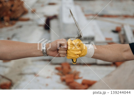Conception of teamwork, success and unity. Side view of two hands of builders on construction site that are knocking the fists Conception of teamwork, success and unity. Side view of two hands of builders on construction site that are knocking the fists 139244044