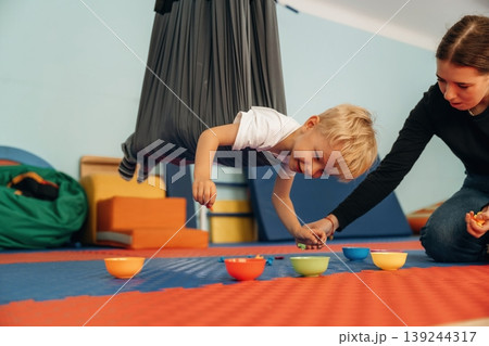 Putting the objects into bowls while hanging. Preschool child with teacher during developmental activities in a playroom Putting the objects into bowls while hanging. Preschool child with teacher during developmental activities in a playroom 139244317