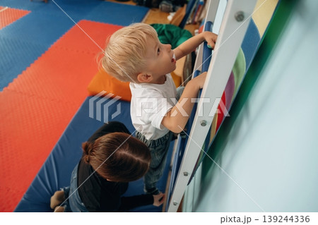 Climbing the ladder that is on the wall. Preschool child with teacher during developmental activities in a playroom 139244336