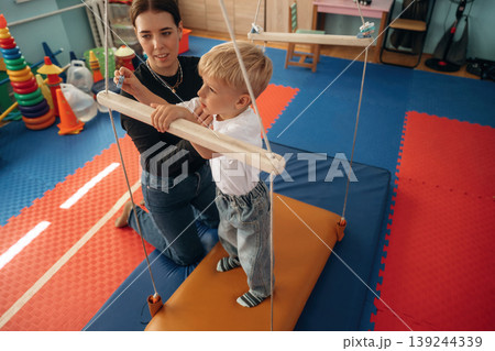 Holding by the wooden stick on surface that is swinging. Preschool child with teacher during developmental activities in a playroom 139244339
