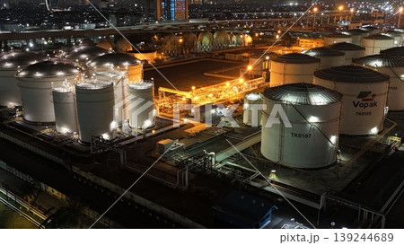 High-angle night view of a fuel storage tank terminal with multiple large, illuminated cylinders and a complex piping infrastructure at an industrial port facility, glowing in the dark. High-angle night view of a fuel storage tank terminal with multiple large, illuminated cylinders and a complex piping infrastructure at an industrial port facility, glowing in the dark. 139244689