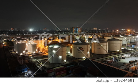 High-angle night view of a fuel storage tank terminal with multiple large, illuminated cylinders and a complex piping infrastructure at an industrial port facility, glowing in the dark. High-angle night view of a fuel storage tank terminal with multiple large, illuminated cylinders and a complex piping infrastructure at an industrial port facility, glowing in the dark. 139244691