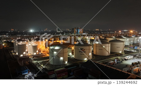 High-angle night view of a fuel storage tank terminal with multiple large, illuminated cylinders and a complex piping infrastructure at an industrial port facility, glowing in the dark. High-angle night view of a fuel storage tank terminal with multiple large, illuminated cylinders and a complex piping infrastructure at an industrial port facility, glowing in the dark. 139244692